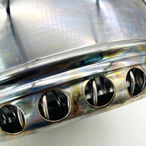 May include: A polished stainless steel cylindrical component featuring a series of circular vents along the lower edge. The metal surface displays iridescent heat-tinted discolouration, and internal coiled wire elements are visible through the openings.