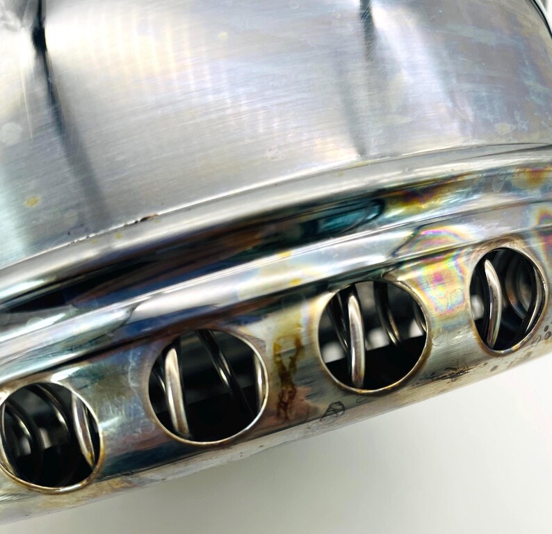 May include: A polished stainless steel cylindrical component featuring a series of circular vents along the lower edge. The metal surface displays iridescent heat-tinted discolouration, and internal coiled wire elements are visible through the openings.