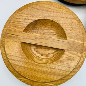 May include: Close-up of a wooden lid with a circular design. The lid is made of light brown wood with a natural grain pattern. It features a recessed area with a small, rectangular handle. The lid appears to be part of a larger set.