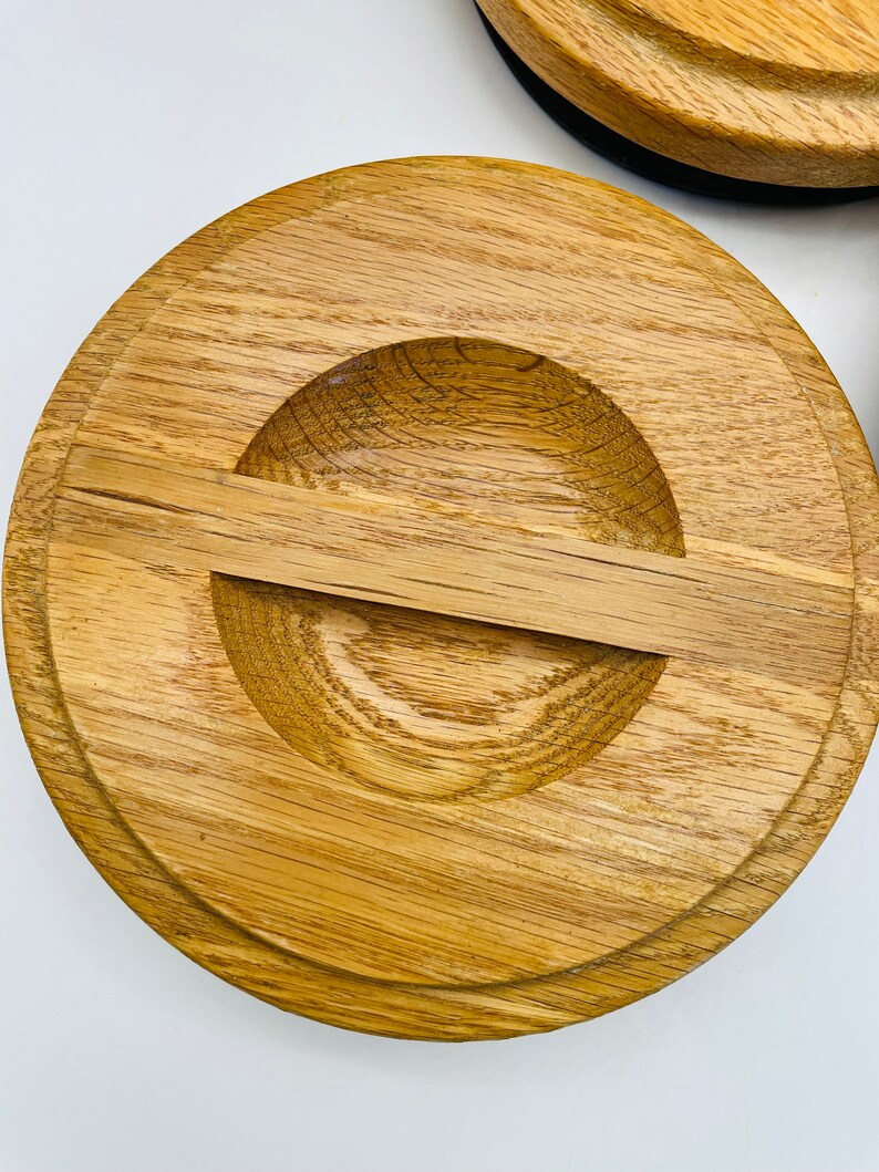 May include: Close-up of a wooden lid with a circular design. The lid is made of light brown wood with a natural grain pattern. It features a recessed area with a small, rectangular handle. The lid appears to be part of a larger set.