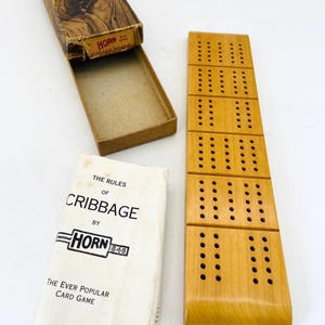 May include: A light brown wooden cribbage board with a grid of holes, accompanied by its original cardboard box featuring a wood grain pattern and a white instruction booklet titled The Rules of Cribbage by Horn 1846, Copyright 1937.