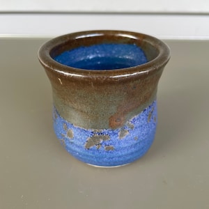 May include: A handmade ceramic cup with a blue interior and a textured exterior. The cup features a gradient of colors, transitioning from a deep blue at the base to a brown rim. The design includes speckled details.