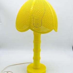 May include: A bright yellow table lamp featuring a dome-shaped shade with a woven grid pattern and a scalloped edge. The lamp has a cylindrical base with horizontal ring details and a smooth circular stand, accompanied by a white power cord.