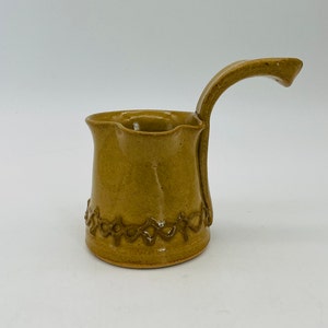May include: A small, hand-thrown ceramic pitcher with a brown glaze. The pitcher has a unique, wavy rim and a decorative band around the bottom. The handle is curved and attached to the side of the pitcher.