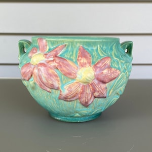 May include: A turquoise ceramic planter with a round shape, two handles, and embossed floral patterns. The flowers are pink with yellow centres. This decorative planter, suitable for indoor or outdoor use, measures approximately 20 cm in diameter.