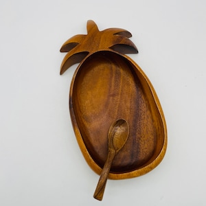 May include: A pineapple-shaped wooden bowl with a smooth, polished finish and visible wood grain patterns. The bowl features a carved leaf design at the top and includes a small, matching wooden spoon resting inside the basin of the bowl.