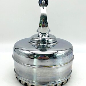 May include: A polished chrome stovetop kettle with a cylindrical body and a vertical handle. The base features a series of circular ventilation holes around the perimeter. The reflective metallic surface shows subtle signs of wear and oxidation.