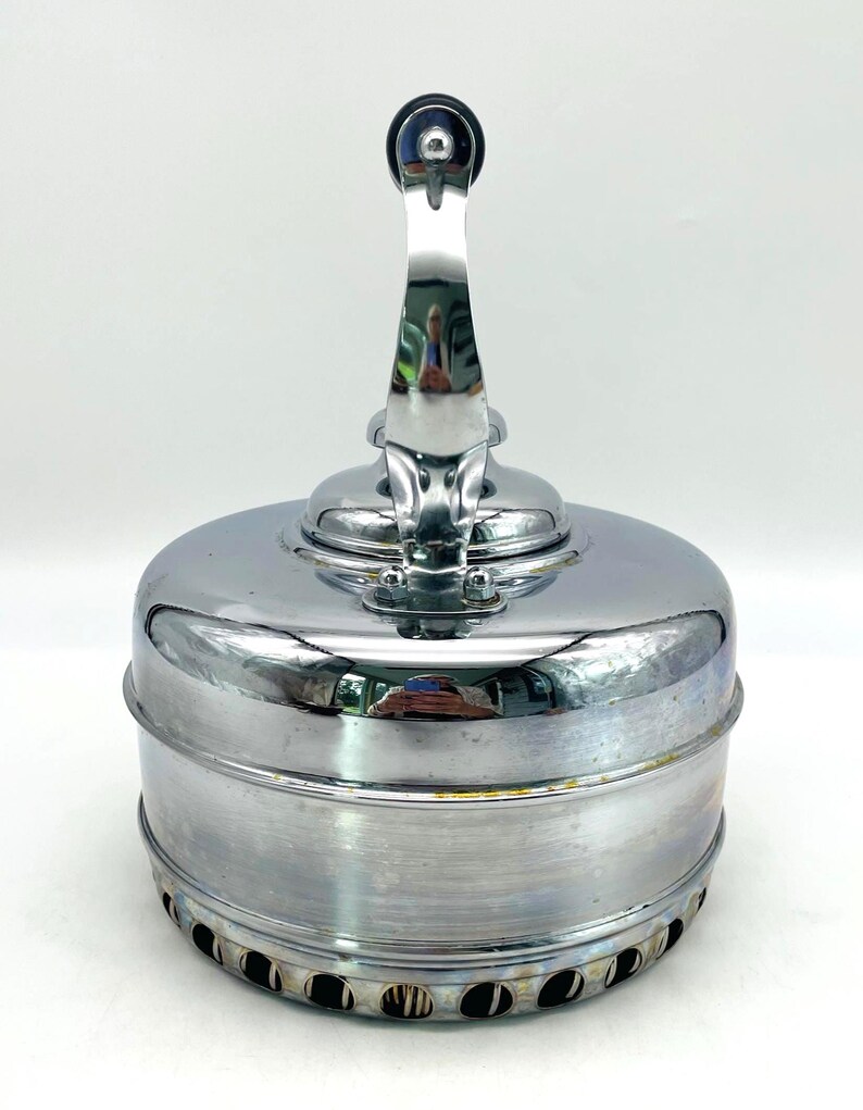 May include: A polished chrome stovetop kettle with a cylindrical body and a vertical handle. The base features a series of circular ventilation holes around the perimeter. The reflective metallic surface shows subtle signs of wear and oxidation.
