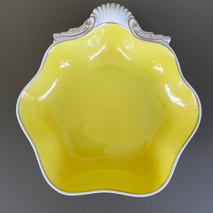 May include: A decorative yellow ceramic bowl with a scalloped edge and a white and gold trim. The bowl has a unique shape and a glossy finish, perfect for serving or display.