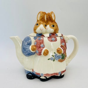 May include: A ceramic teapot featuring a whimsical design of two rabbits. The teapot is primarily cream-colored, with details in brown, blue, and red. The lid is topped with two rabbit heads, and the teapot has a handle and spout.