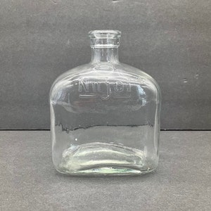 May include: A clear glass bottle with a rectangular body and a short, narrow neck. The word Nujol is embossed in capital letters on the front surface of the glass. The bottle features a simple, functional design with a smooth, transparent finish.