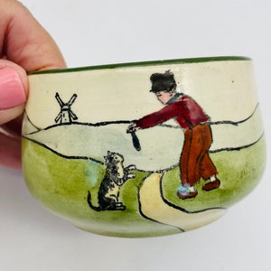 May include: A small, hand-painted ceramic bowl with a whimsical scene. The bowl features a figure in a red jacket and brown pants, a black cat, and a windmill. The rim is green, and the background is a light cream color.