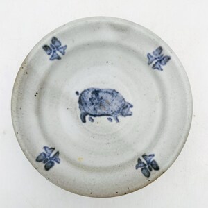 May include: A round, off-white ceramic plate with a speckled texture. The center features a blue hand-painted pig illustration. Four small, blue, butterfly-like floral motifs are evenly spaced around the rim of the plate.