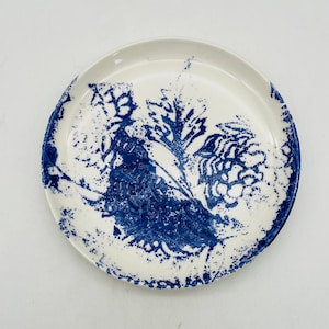 May include: White ceramic plate with a blue floral design. The plate features a detailed, hand-painted pattern of leaves and flowers, with a textured, speckled appearance. The rim of the plate is slightly raised.