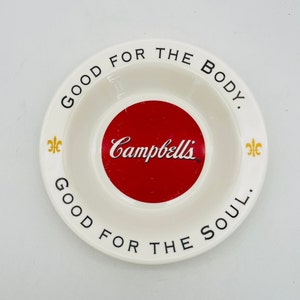 May include: A white ceramic bowl with the Campbell's logo in the center, featuring a red circle with the brand name in white script. The bowl has the text "GOOD FOR THE BODY" and "GOOD FOR THE SOUL" around the rim, with gold fleur-de-lis accents.