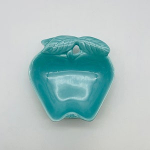 May include: A turquoise ceramic apple-shaped dish with two leaves at the top. The dish has a smooth, glossy finish and a small hole in the center of the leaves. This decorative dish is ideal for serving snacks or as a decorative accent.
