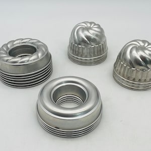 May include: A collection of four vintage aluminum Bundt cake molds in various shapes. The molds include a ring shape and three dome-shaped molds with decorative fluted and twisted designs. The molds are made of shiny, silver-colored metal.