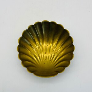 May include: A small, decorative, golden-colored seashell-shaped dish. The dish has a scalloped edge and a textured surface, with a gradient of color from light gold to a darker, more aged gold.