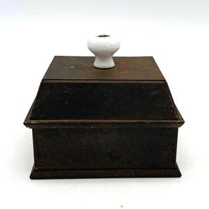 May include: A square, dark brown, weathered metal box with a tapered lid and a smooth, white, rounded ceramic knob centered on top. The metal surface shows signs of oxidation and age, featuring a flat base and a distinct, stepped lid design.