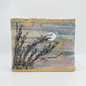 May include: A rectangular ceramic vase with a textured, sandy-colored rim and base. The surface features a painted landscape with a pale blue and pink sky, a white circular moon, and dark, spindly plant silhouettes in the foreground.