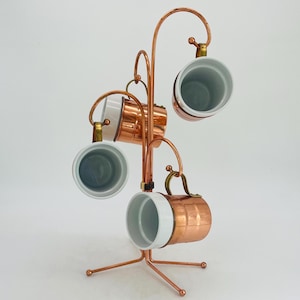 Może przedstawiać: Miedziany stojak na kubki z pięcioma kubkami. Kubki mają miedziany wygląd zewnętrzny i białe wnętrze. Stojak ma zakrzywioną konstrukcję z okrągłą podstawą. Kubki są zawieszone na stojaku za uchwyty.
