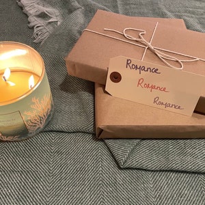 Puede incluir: Dos paquetes envueltos con un exterior de papel marr&oacute;n y una cuerda atada alrededor. Los paquetes est&aacute;n etiquetados con "Romance" en escritura a mano. Una vela encendida con un dise&ntilde;o verde y naranja est&aacute; en primer plano.