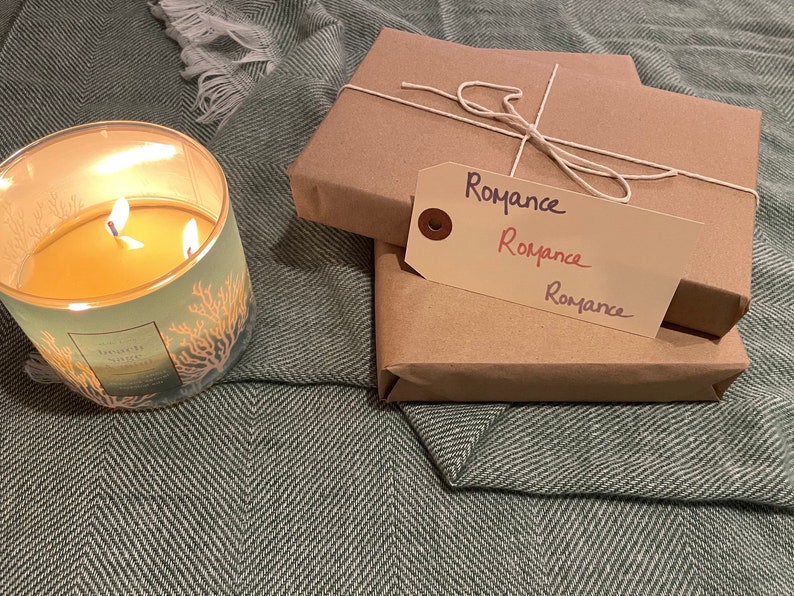 Puede incluir: Dos paquetes envueltos con un exterior de papel marr&oacute;n y una cuerda atada alrededor. Los paquetes est&aacute;n etiquetados con "Romance" en escritura a mano. Una vela encendida con un dise&ntilde;o verde y naranja est&aacute; en primer plano.