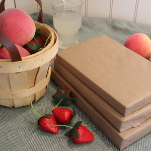 Puede incluir: Una pila de tres libros envueltos en papel marr&oacute;n sobre una superficie con estampado verde y blanco. Una cesta llena de fresas rojas y melocotones rosas se encuentra junto a los libros.