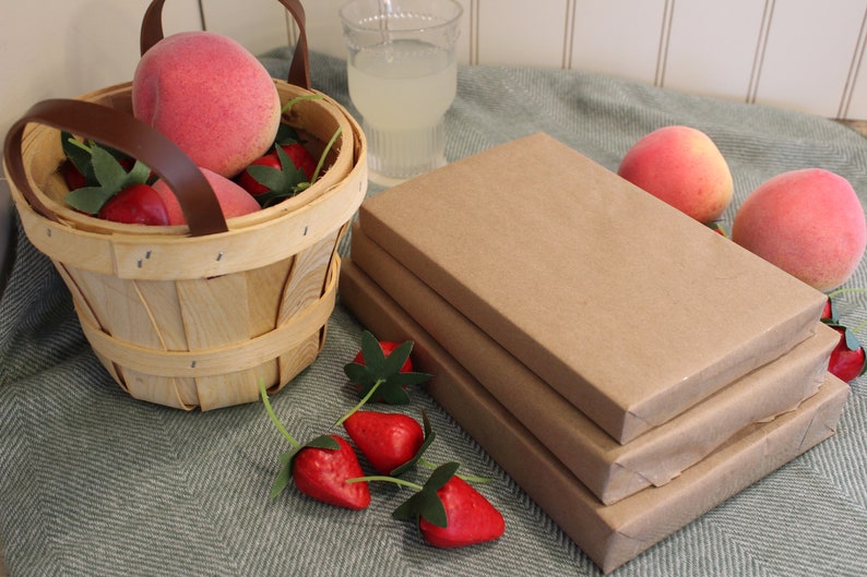 Puede incluir: Una pila de tres libros envueltos en papel marr&oacute;n sobre una superficie con estampado verde y blanco. Una cesta llena de fresas rojas y melocotones rosas se encuentra junto a los libros.