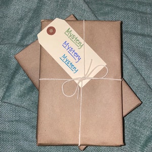 Puede incluir: Dos paquetes envueltos en papel marr&oacute;n, atados con cuerda y una etiqueta de regalo blanca que dice "Misterio" en verde, azul y morado.