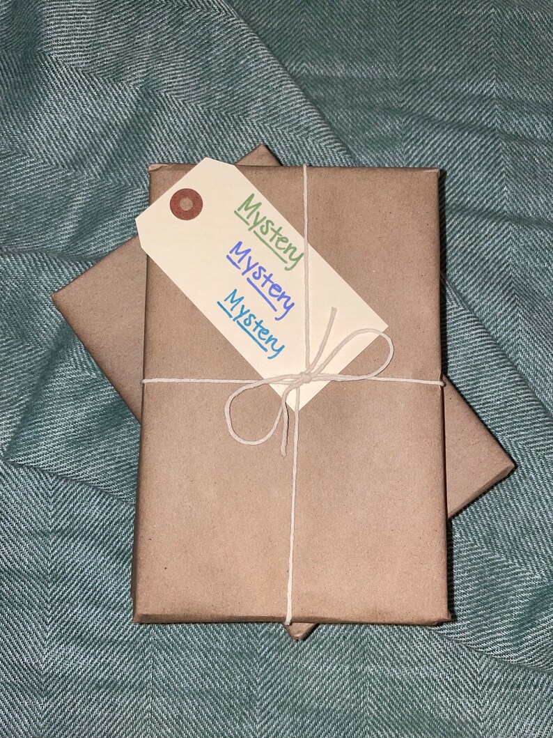 Puede incluir: Dos paquetes envueltos en papel marr&oacute;n, atados con cuerda y una etiqueta de regalo blanca que dice "Misterio" en verde, azul y morado.