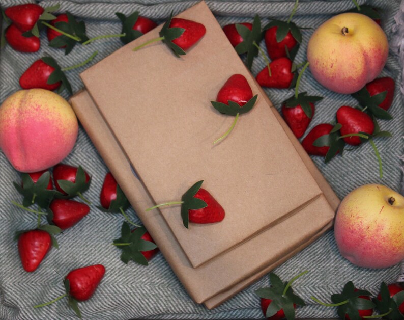 Puede incluir: Dos cuadernos marrones apilados uno encima del otro sobre un fondo de tela rayada verde y blanca. Los cuadernos est&aacute;n rodeados de fresas rojas y fruta de color melocot&oacute;n.