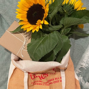Puede incluir: Una bolsa de lona marr&oacute;n con el texto "The Tines Club" y "Fueling good vibes all day" impreso en rojo. La bolsa est&aacute; llena de un ramo de girasoles y un regalo envuelto en papel marr&oacute;n.