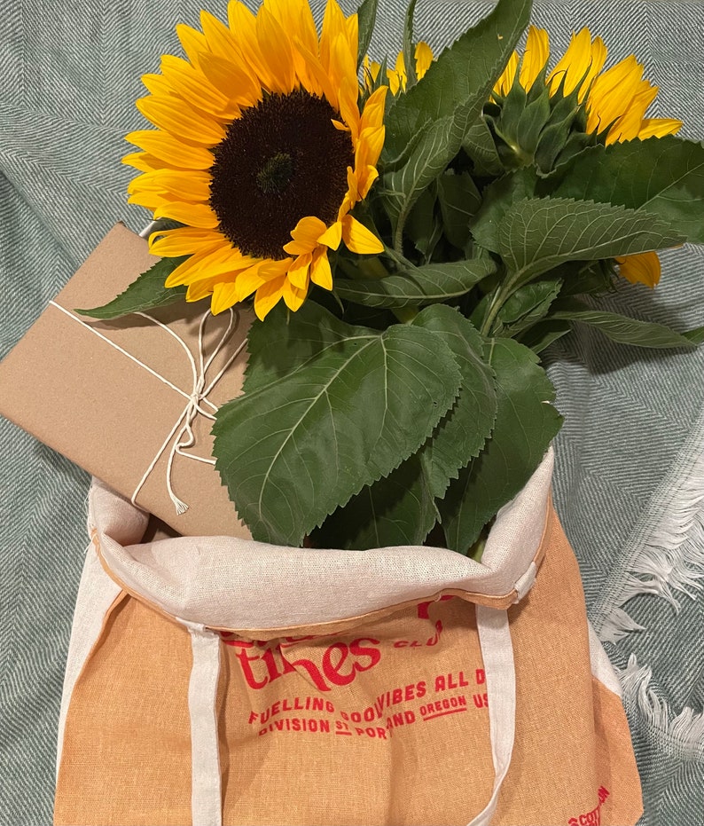 Puede incluir: Una bolsa de lona marr&oacute;n con el texto "The Tines Club" y "Fueling good vibes all day" impreso en rojo. La bolsa est&aacute; llena de un ramo de girasoles y un regalo envuelto en papel marr&oacute;n.