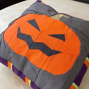 Könnte beinhalten: Quadratisches Halloween-Kissen mit einem orangefarbenen Kürbisgesicht auf grauem Hintergrund. Das Kissen hat einen lila Rand mit orangefarbenen und grünen Streifen. Das Design ist im Patchwork-Stil.