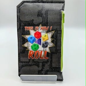 May include: A rectangular black fabric pouch featuring a graphic of multi-colored polyhedral dice and the text "THIS IS HOW I ROLL" in a comic-style burst. The pouch has a bright lime green zipper closure along the right side and a dark pattern.