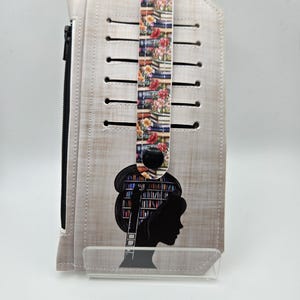May include: A light beige wallet with a black zipper and a floral strap. The wallet features a silhouette of a woman with a bookshelf design. The strap is decorated with images of books and flowers.