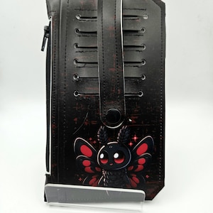 May include: Black wallet with a red and black moth illustration. The wallet has a zipper closure, a handle, and a button clasp. The moth has red wings and a cute expression. The wallet is made of a dark material with a distressed look.