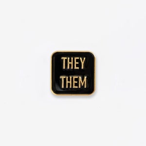May include: Black square pin with gold trim and the text "THEY THEM" in gold letters.