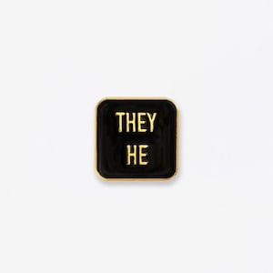 May include: Black enamel pin with gold trim featuring the words "THEY HE" in gold lettering.