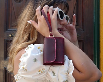 May include: A woman holds a burgundy leather wallet with a pink stitch detail and a gold clasp. The wallet has a strap and is hanging from her hand.