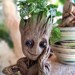 Puede incluir: Un macetero de cerámica marrón y verde con forma del personaje Groot de la película Guardianes de la Galaxia. El macetero tiene una cara sonriente y sostiene una planta con hojas verdes.