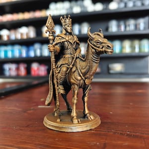 May include: A bronze-colored figurine of a king riding a camel, holding a staff. The king wears a crown and ornate clothing. The figurine is on a round base, with detailed textures. The background is blurred, showing shelves with various items.