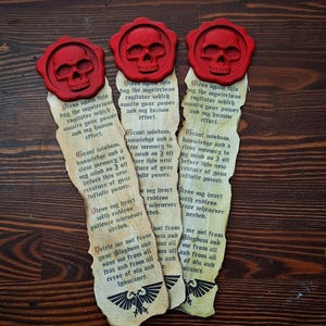 Puede incluir: Tres marcapáginas hechos a mano con temática gótica. Cada marcapáginas presenta un sello de cera rojo con forma de calavera en la parte superior y papel envejecido con texto impreso. La parte inferior de cada marcapáginas tiene un emblema en blanco y negro.