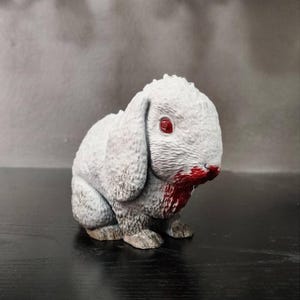 Op de afbeelding: Een wit konijnenbeeldje met rode ogen en bloed dat van zijn neus en mond druipt. Het beeldje staat op een zwarte ondergrond.