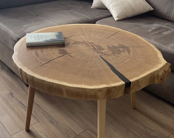 May include: A round wooden coffee table with a natural wood finish and a black epoxy resin fill. The table has four thin, light wood legs and is sitting on a wood floor. A book is resting on the table.