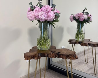 May include: A wooden table with a live edge top and gold hairpin legs. A vase with pink peonies and eucalyptus is on the table.