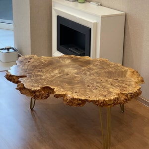 Könnte beinhalten: Ein großer, ovaler Couchtisch aus einer natürlichen Holzplatte mit einem einzigartigen, gemaserten Muster. Der Tisch hat eine lebende Kante und wird von vier goldenen Haarnadelbeinen getragen.