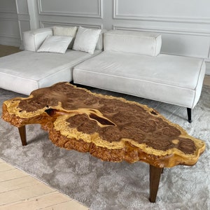May include: A large, organic-shaped coffee table crafted from a single piece of wood with a natural, brown and yellow wood grain. The table has a unique, irregular shape and is supported by four dark brown legs.