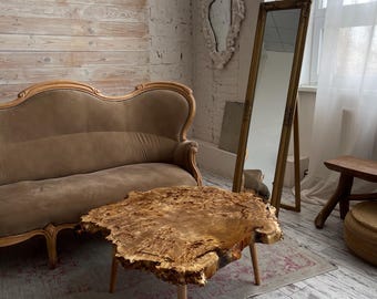 May include: A rustic wooden coffee table with a live edge top and natural wood grain. The table has four slender legs and is sitting on a rug with a floral pattern.